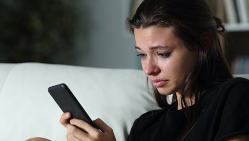Foto einer weinenden jungen Frau auf der Couch, die auf ihr Smartphone schaut